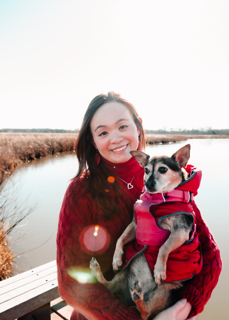 Photo of a smiling Sharon carrying her chihuahua-mix furkid in front of a lake in winter.